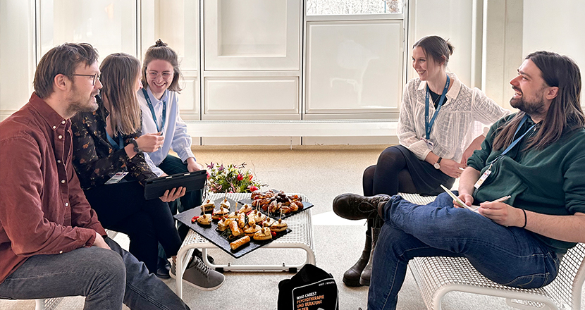 Fünf Personen sitzen in einem hellen Raum um einen kleinen Tisch mit Snacks und Getränken; sie unterhalten sich in entspannter Atmosphäre